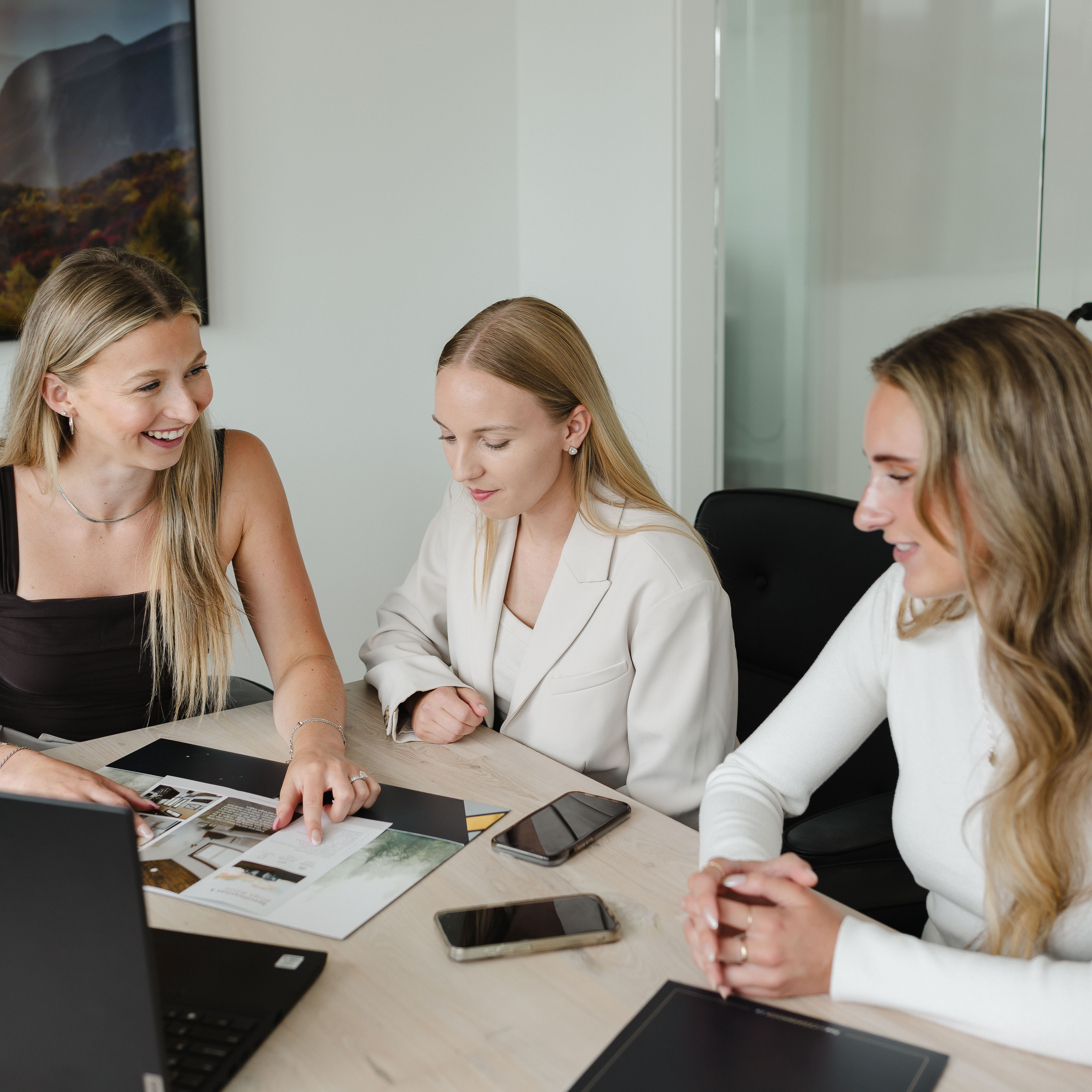 Three women collaborating at a desk with a laptop and smartphone, smiling and engaged.