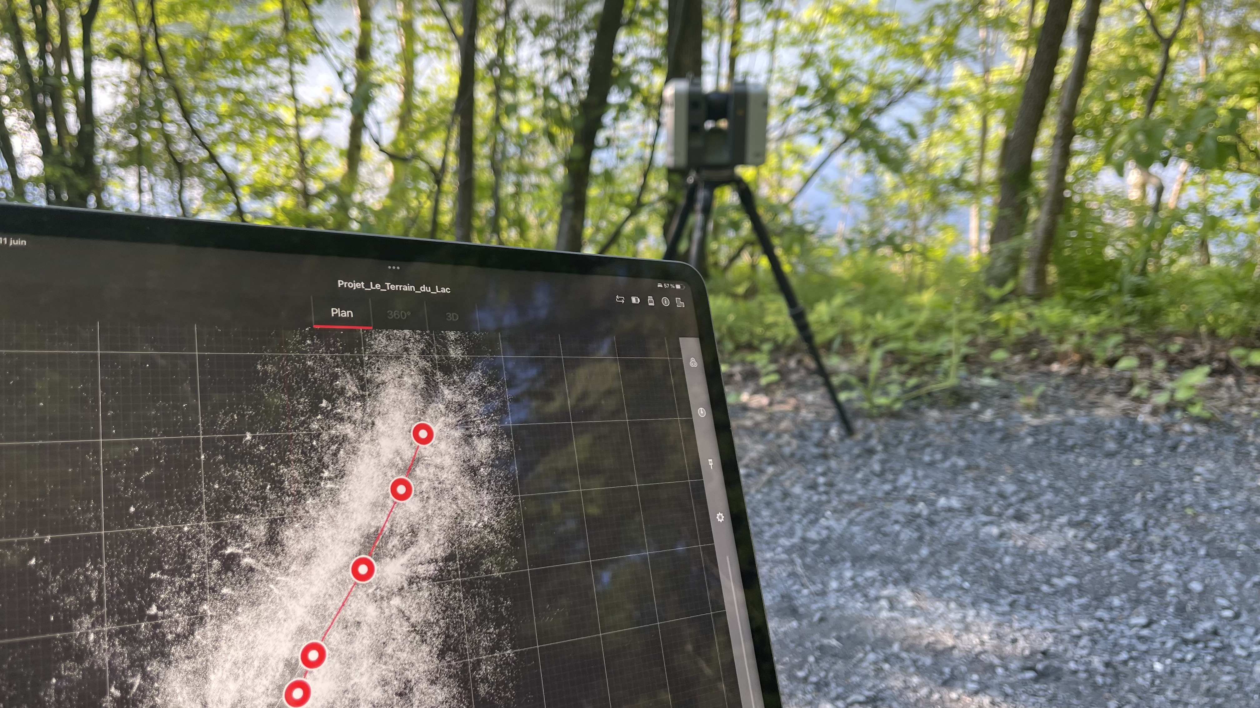 Tablette affichant un projet de carte topographique avec plusieurs points connectés, dans un environnement forestier avec un équipement de relevé sur trépied en arrière-plan.