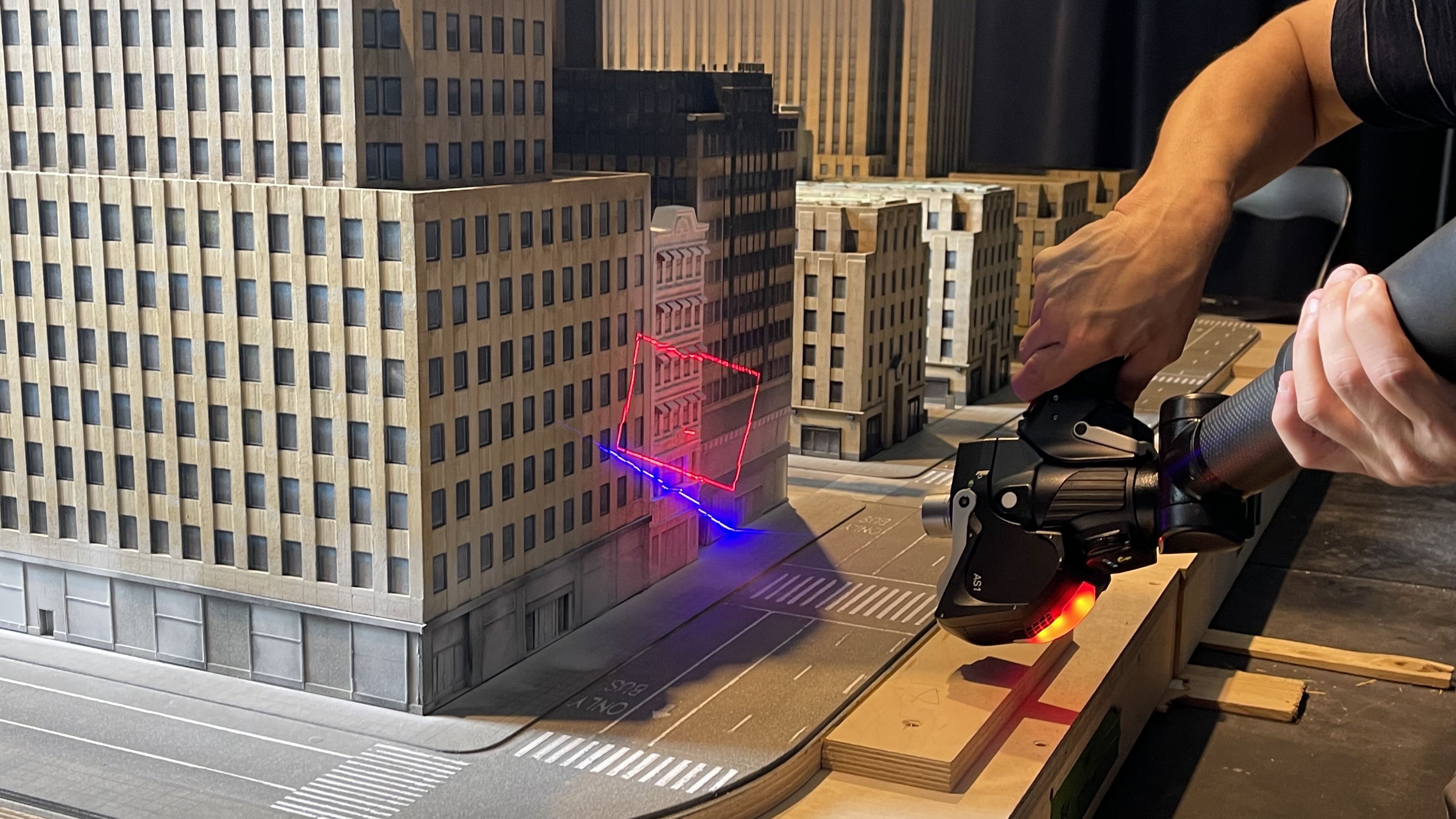 Une maquette de ville avec un faisceau laser rouge et bleu projeté sur un bâtiment, une personne tenant un appareil de balayage 3D.