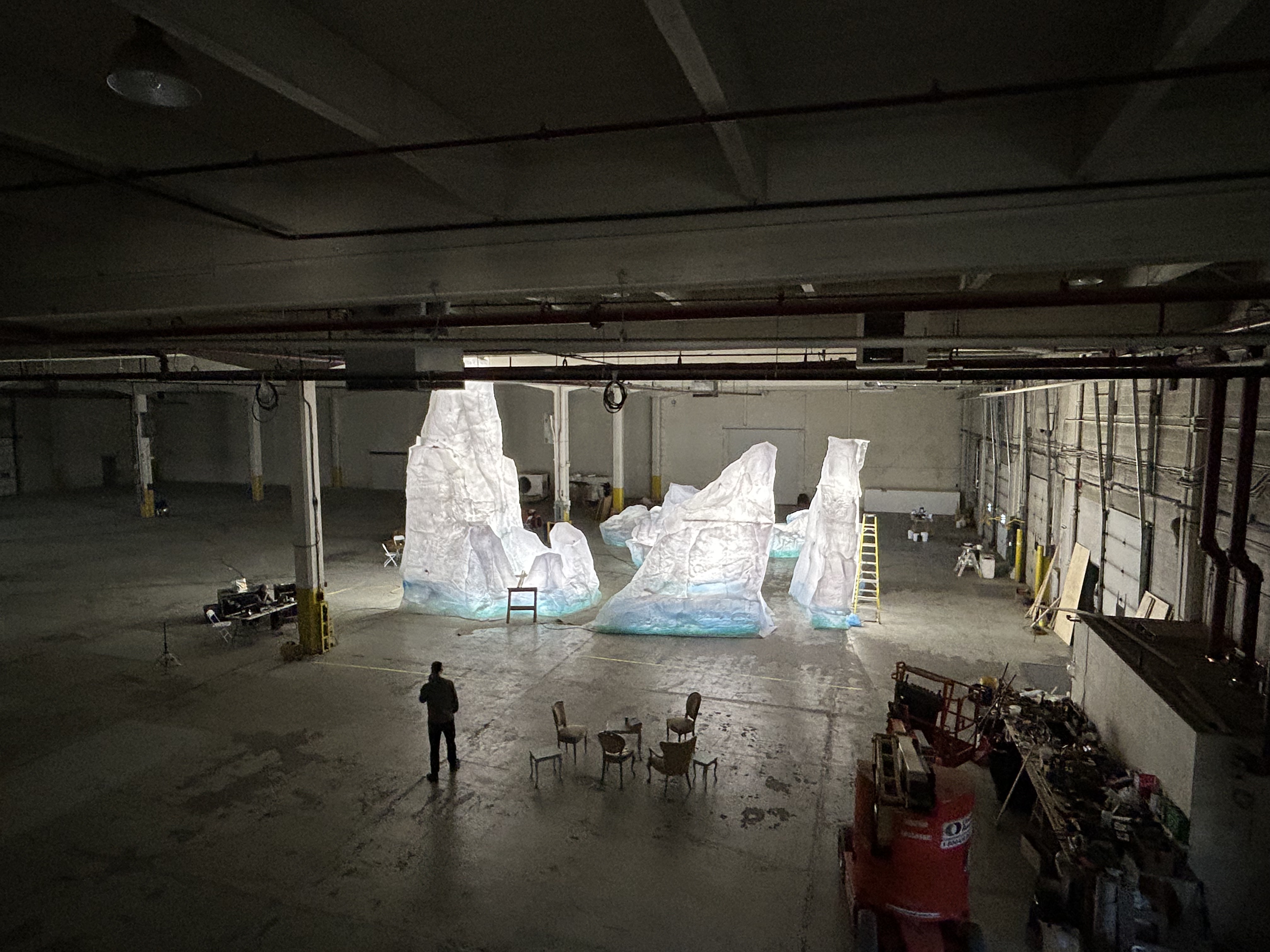 Grand entrepôt avec une installation artistique représentant des grandes formations rocheuses blanches éclairées, avec des chaises et une personne debout à proximité.