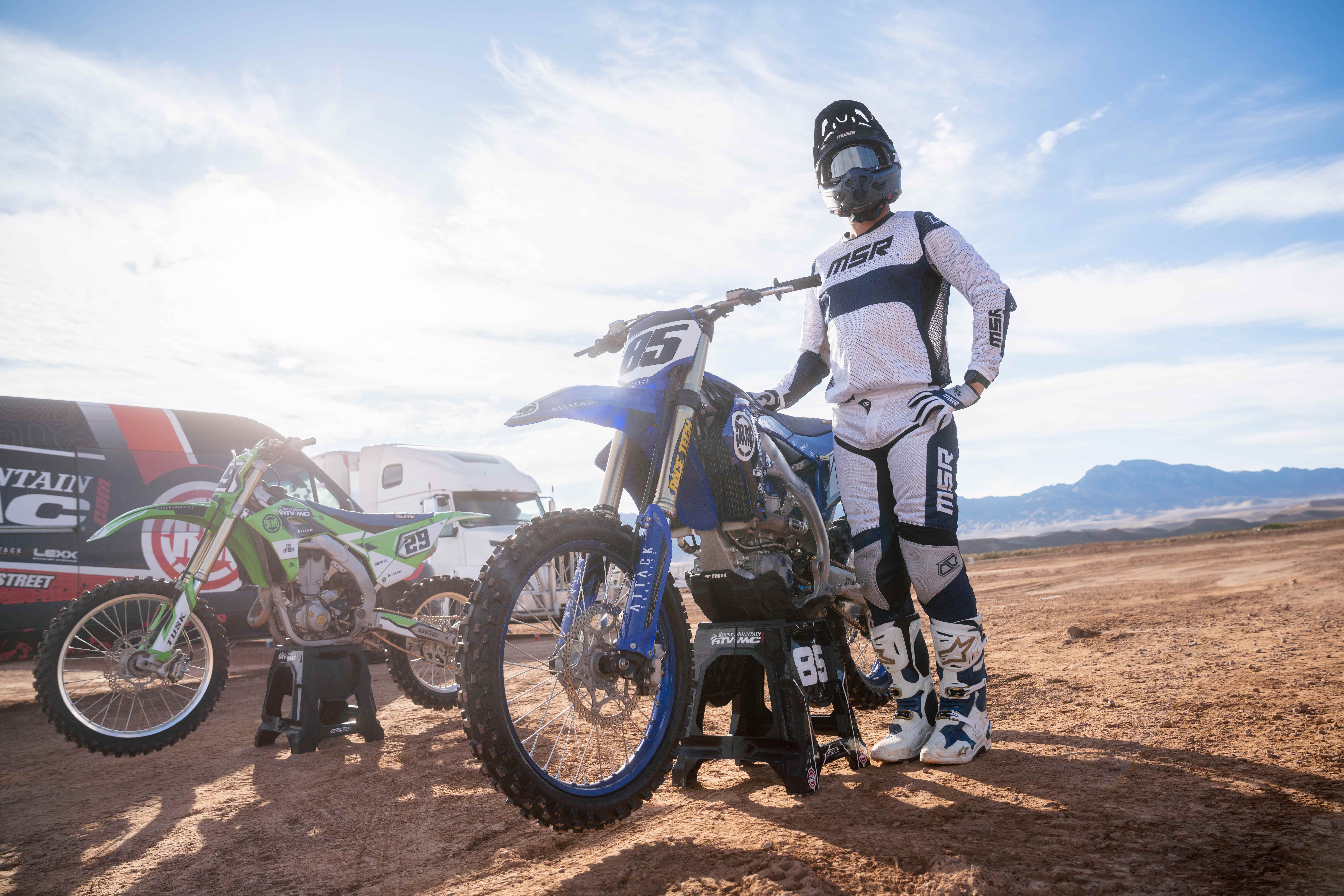 A rider wearing blue and whit gear standing next to his bike.