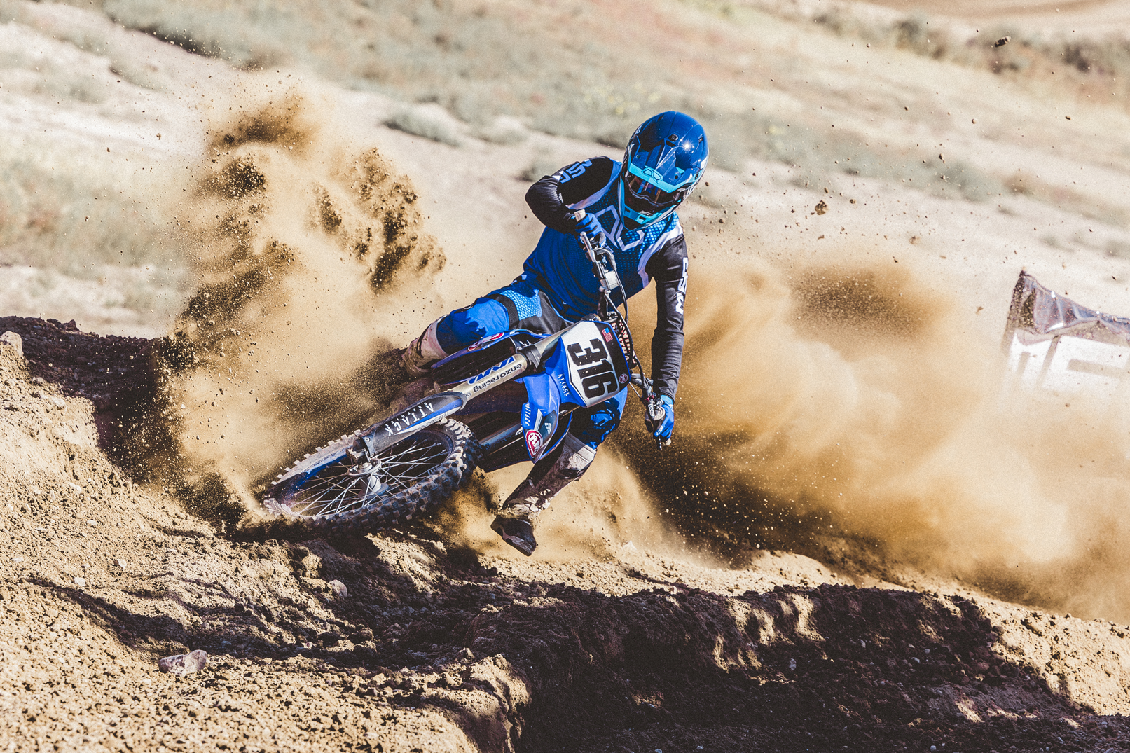 a rider in blue gear taking a turn on a dirt bike and spraying dirt in a could.