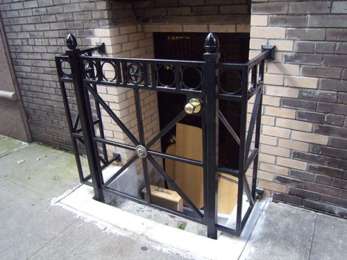 DG161 Ornamental metal gate enclosure features a geometric X-brace design with circular motifs along the upper rail, decorative rosettes at the intersections, and square tube posts capped with acorn finials, surface-mounted to a concrete curb