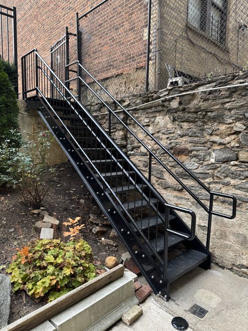 Steel staircase featuring welded tubular handrails, vertical balusters, diamond plate treads, and anchor bolts securing the structure to a concrete base.