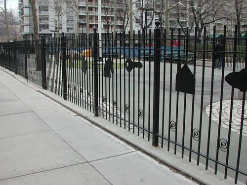 R159 Welded steel perimeter fence featuring vertical square pickets, reinforced double horizontal rails, decorative scroll accents at the base, and custom-cut flat bar silhouette inlays mounted between posts with ball finials, surface-anchored into concrete pavement along a public sidewalk