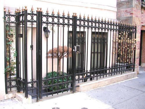 R161 Decorative wrought iron entry fence and gate system featuring vertical square bar pickets with spearpoint finials, double horizontal rails, and a repeating punched-ring motif along both the top and bottom panels, all mounted to steel posts with cast finial caps and set into a masonry curb along the sidewalk.