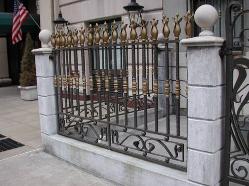 R164 Decorative wrought iron fence section framed between masonry piers, featuring vertical balusters with alternating gold-painted urn finials and turned spindle collars, a mid-rail with ornamental knuckles, and a base panel with scroll and heart-shaped motifs enclosed within a square steel frame.