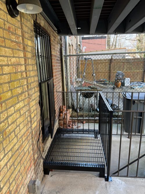 ST032 Welded steel access platform with an open bar grating deck, supported by cantilevered steel angles and framed with square tube guardrails, anchored to a masonry wall and adjacent concrete landing