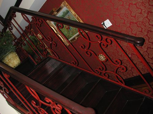 R166 Interior stair railing features hand-forged scrollwork in a symmetrical S-curve and C-curve motif, fabricated from flat and round bar stock, integrated with circular cast medallions, and finished in a high-gloss red enamel beneath a stained wooden handrail with volute termination