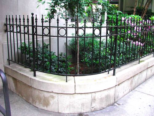 R180 Wrought iron fence features vertical pickets with alternating spear-point and ball finials, circular ring accents set between double horizontal rails, and core-drilled posts mounted into a curved stone planter wall