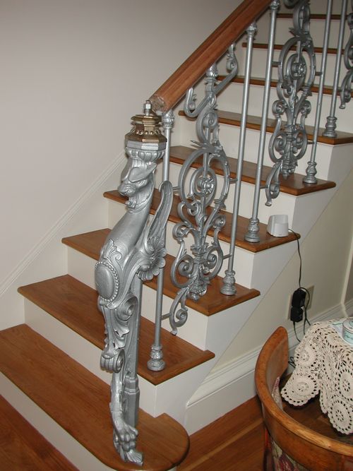 R183 Highly ornamental stair railing features cast iron balusters with scroll and foliate motifs, capped by a stained wood handrail, and anchored by an elaborate cast iron newel post sculpted in the form of a stylized griffin with neoclassical detailing and acanthus leaf accents.