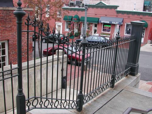 R215 ornamental iron railing features closely spaced vertical pickets topped with decorative spear-point finials and anchored by heavy turned newel posts with urn-style caps. A repeating circular ring motif along the bottom rail