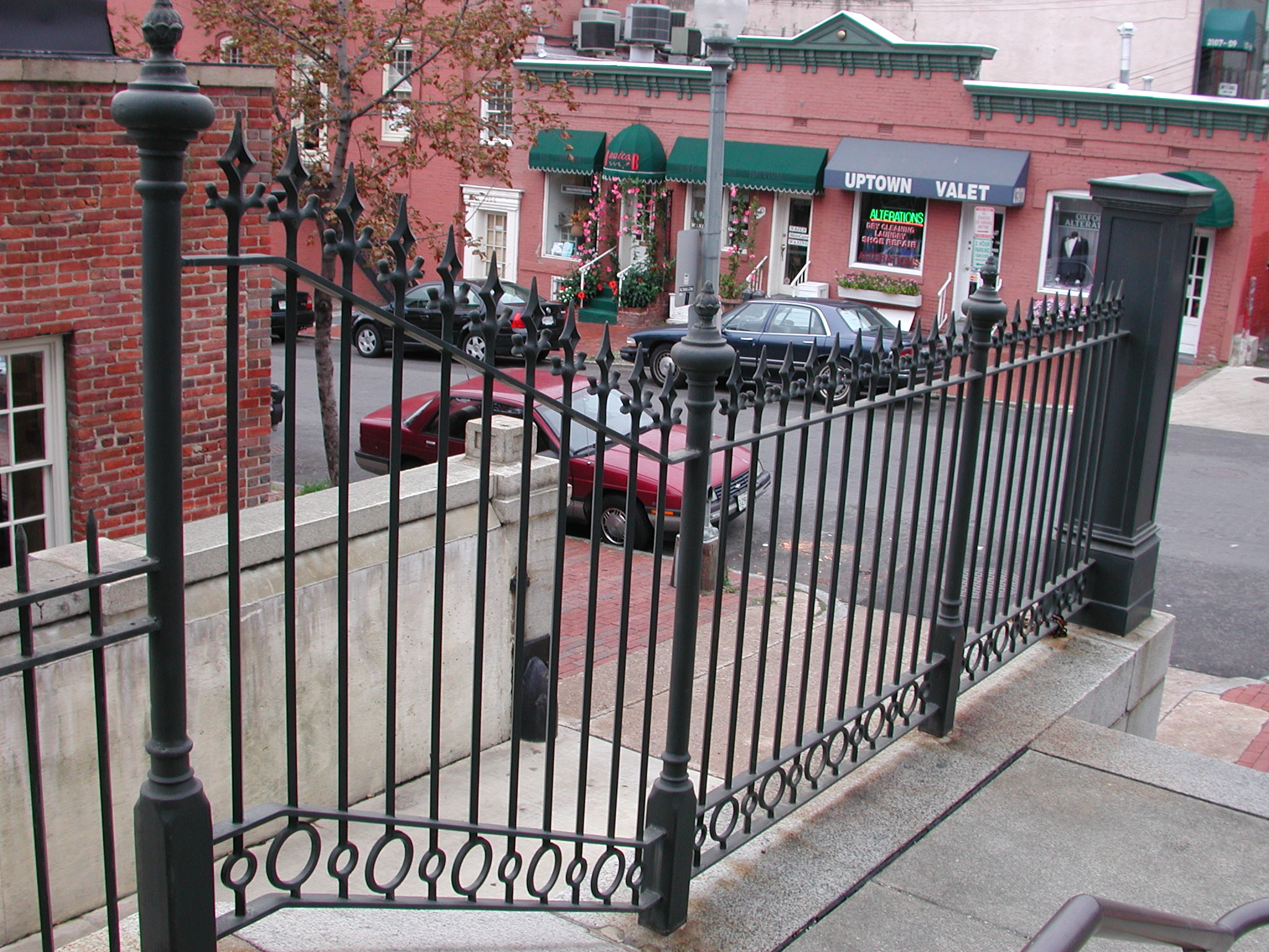 R215 ornamental iron railing features closely spaced vertical pickets topped with decorative spear-point finials and anchored by heavy turned newel posts with urn-style caps. A repeating circular ring motif along the bottom rail