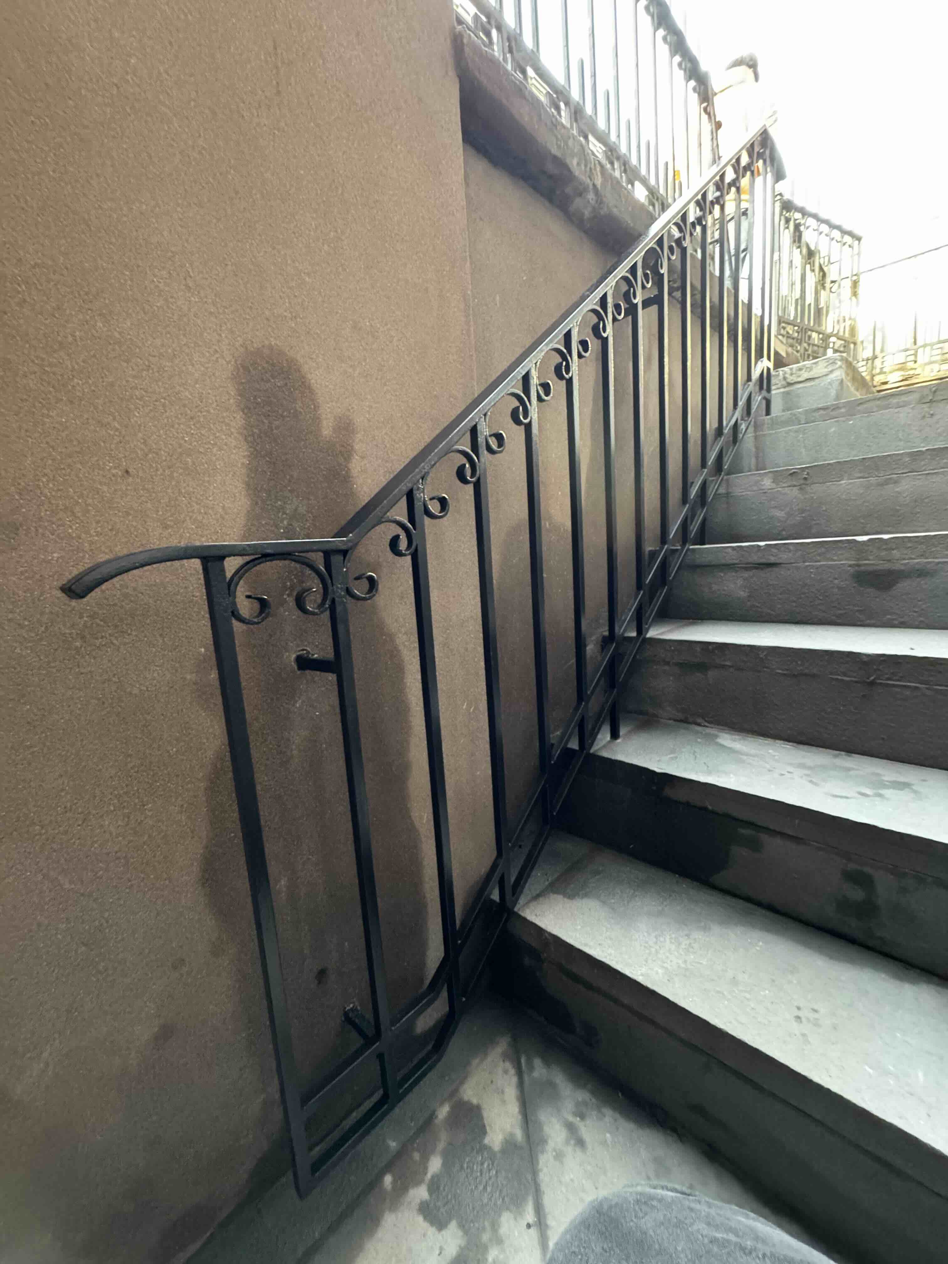 R223 Custom black steel stair railing with vertical pickets, subtle scrollwork accents, and a smooth continuous handrail mounted alongside concrete exterior steps.