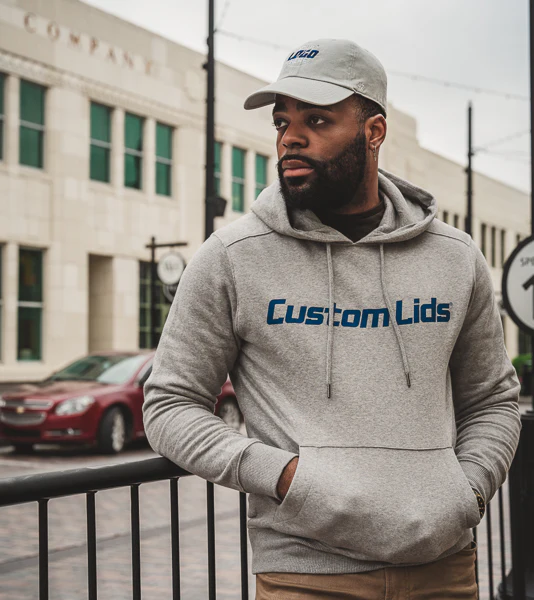 Man wearing a gray Custom Lids branded hoodie and cap standing on a city street.