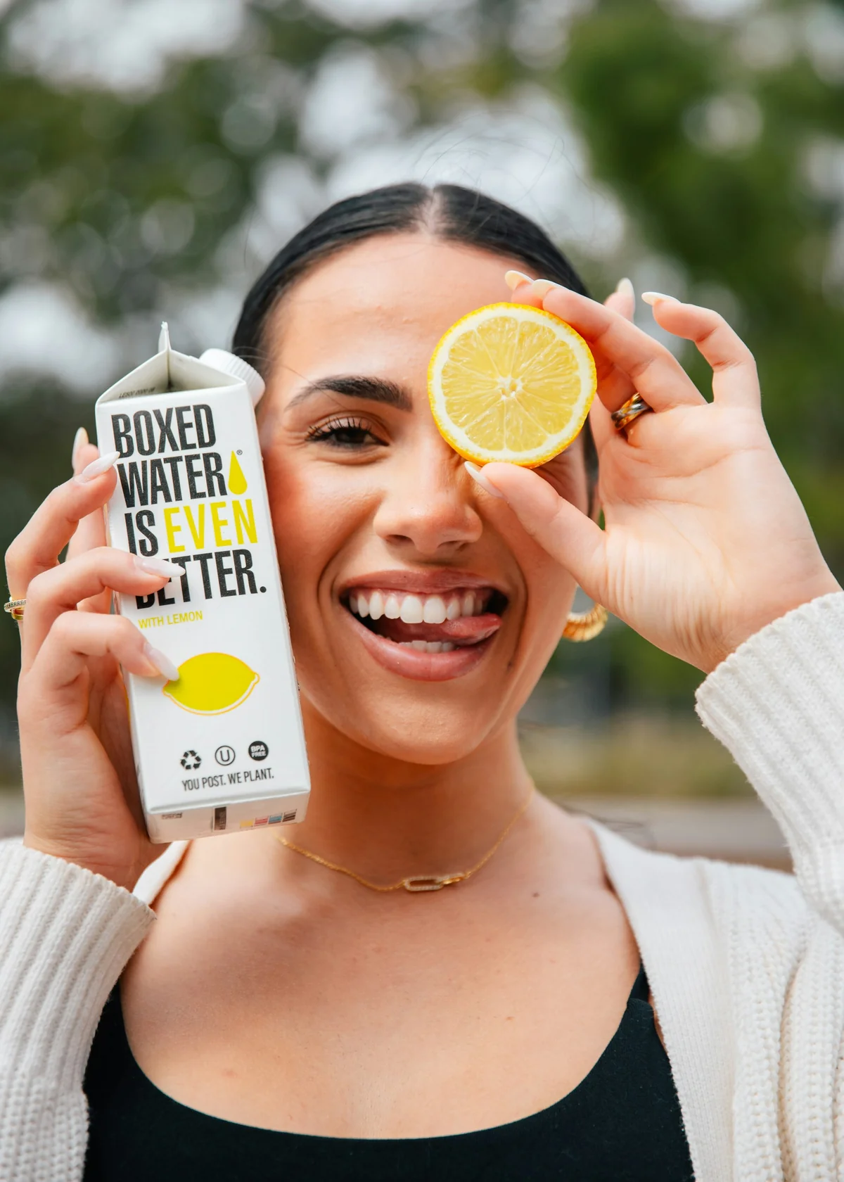 Woman smiling while holding a lemon-flavored Boxed Water carton and a lemon slice over her eye.