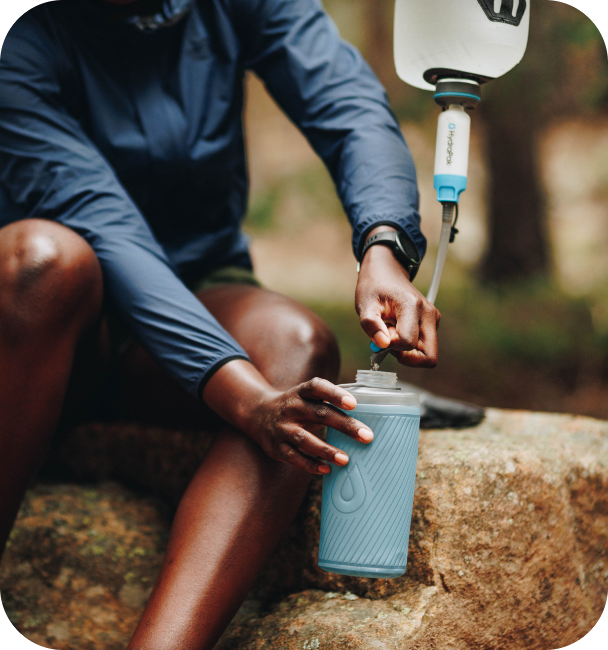 Hiker filling a HydraPak collapsible water flask from a hydration reservoir outdoors on a rock.