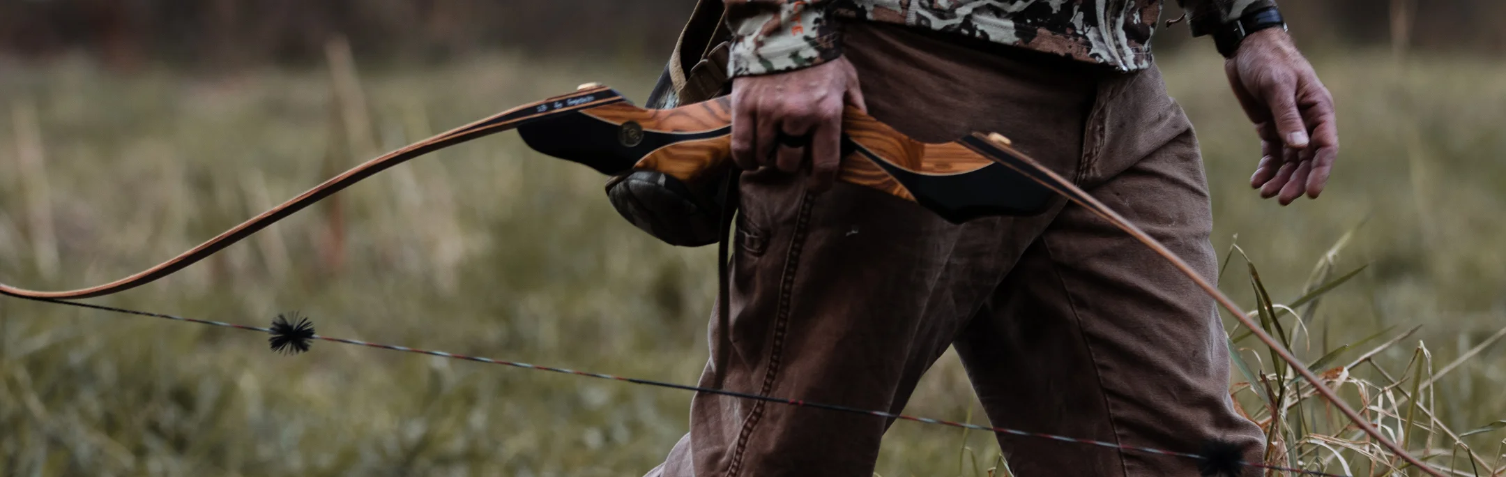 Close-up of a hunter in camouflage gripping a wooden recurve bow in a grassy field