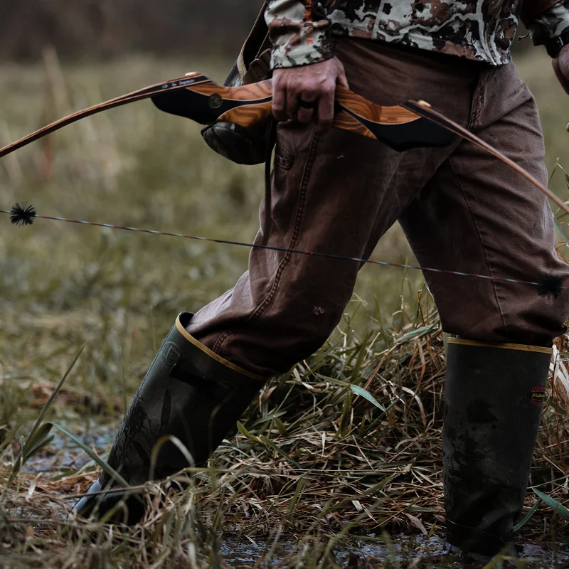 Archer in camouflage carrying a wooden recurve bow while walking through a marshy field