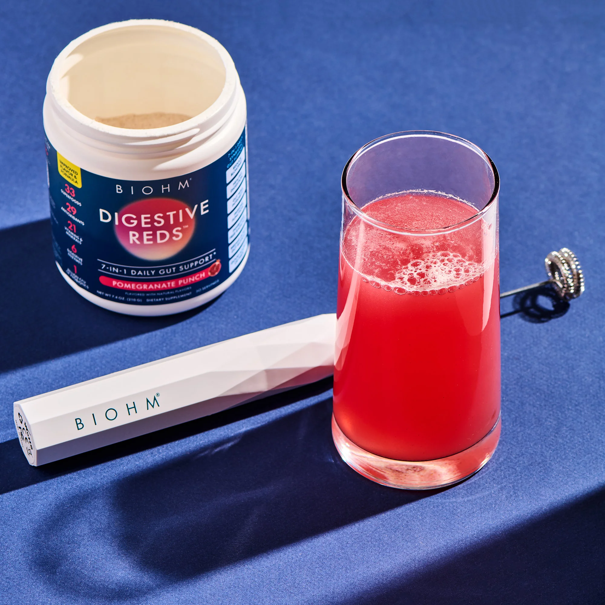 Container of BIOHM Digestive Reds powder, a milk frother, and a glass of mixed pomegranate punch drink.