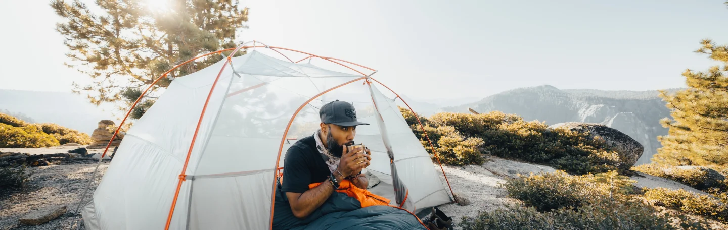 Camper in a Branded Bills hat sipping from a mug inside a tent in the mountains.