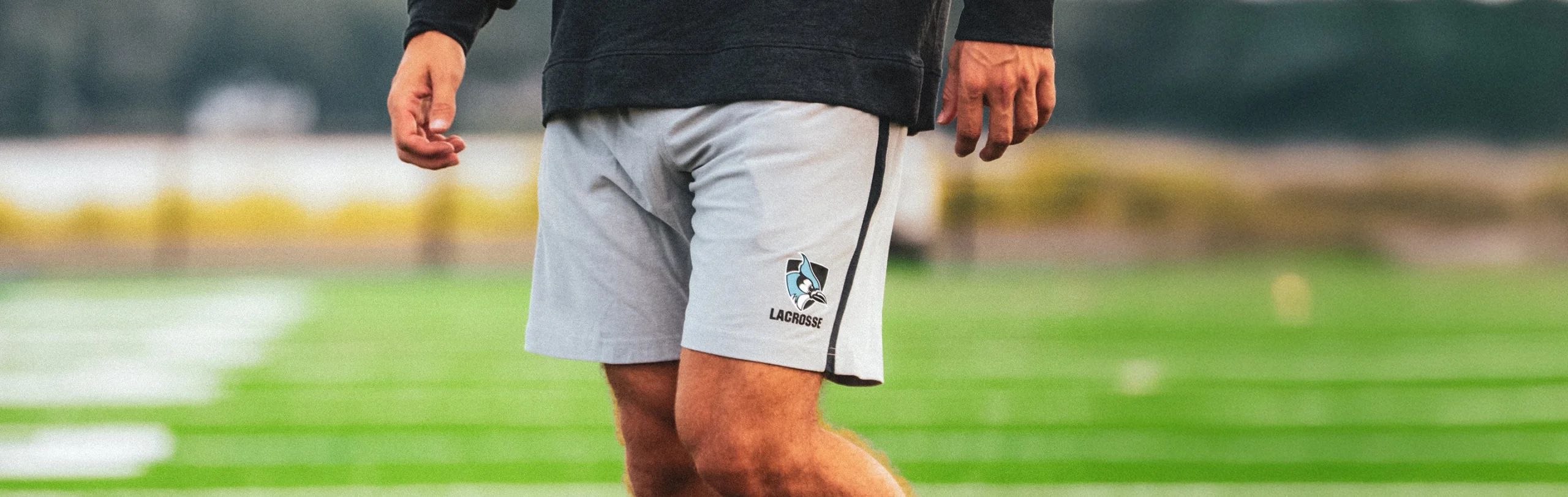Lacrosse player on a turf field wearing gray shorts with a blue jay lacrosse team logo.