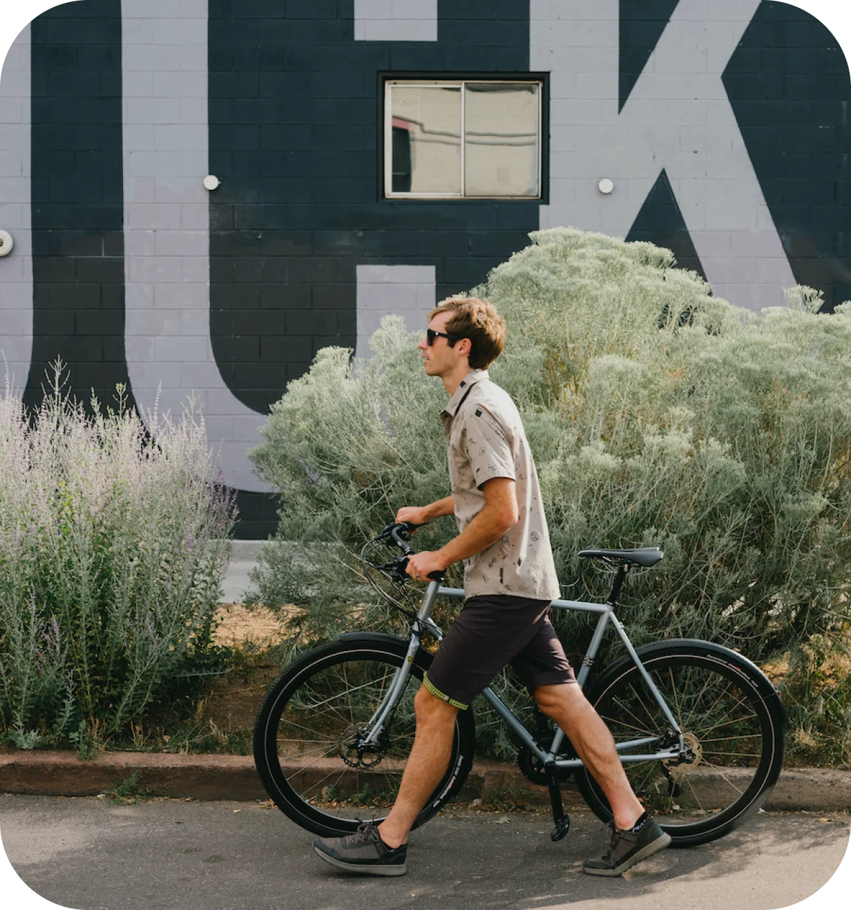 Man in a PEARL iZUMi shirt walking a bike past a mural and sagebrush.