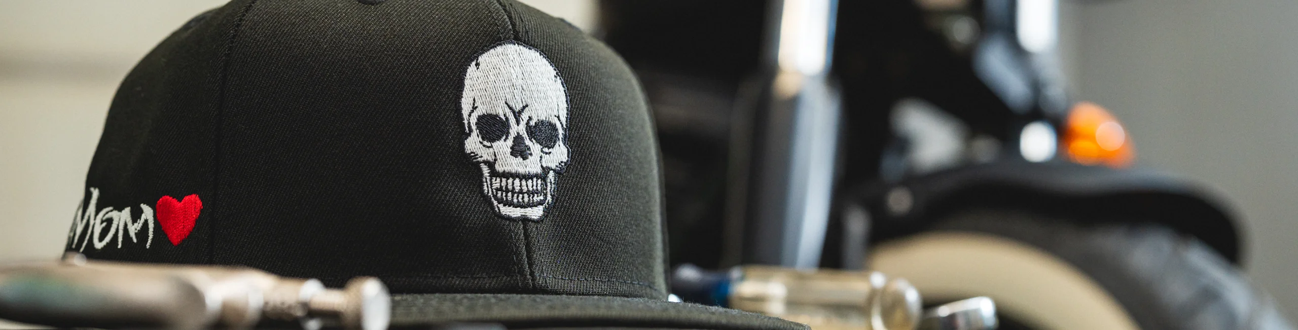 Close-up of a black Custom Lids skull-embroidered snapback with Mom heart stitching beside a motorcycle.