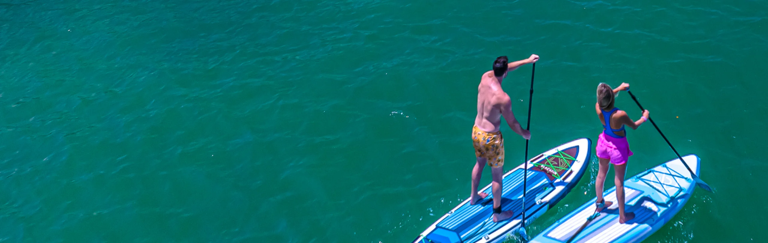 Two people paddling iROCKER stand-up paddle boards across emerald green water.