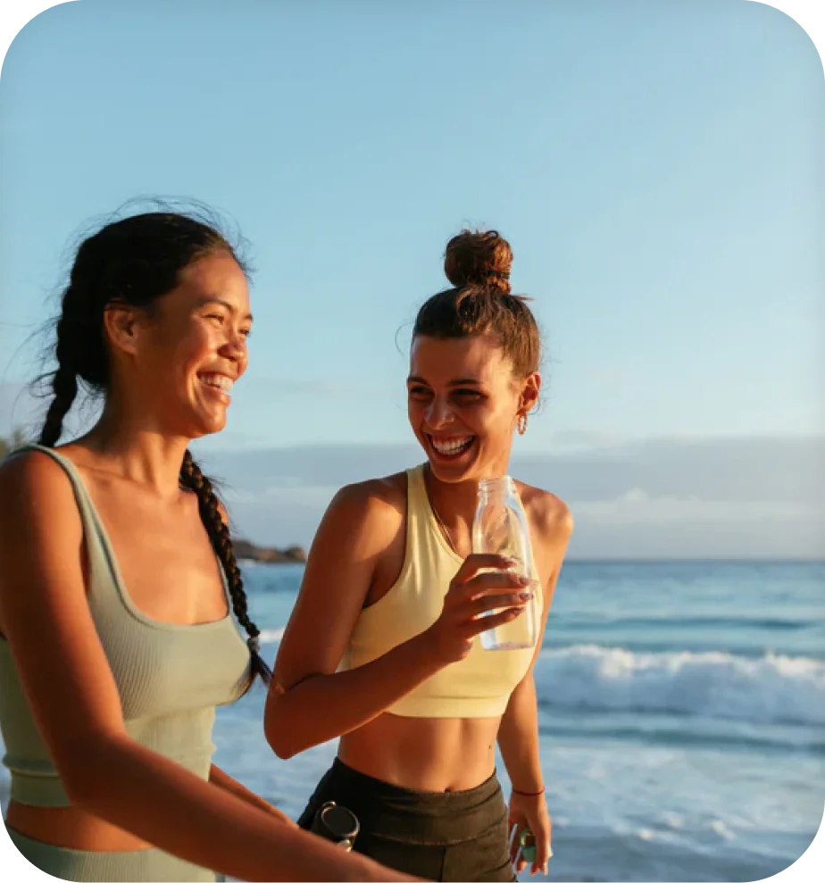 MegaFood lifestyle photo of two women laughing on the beach, one holding a glass supplement bottle.