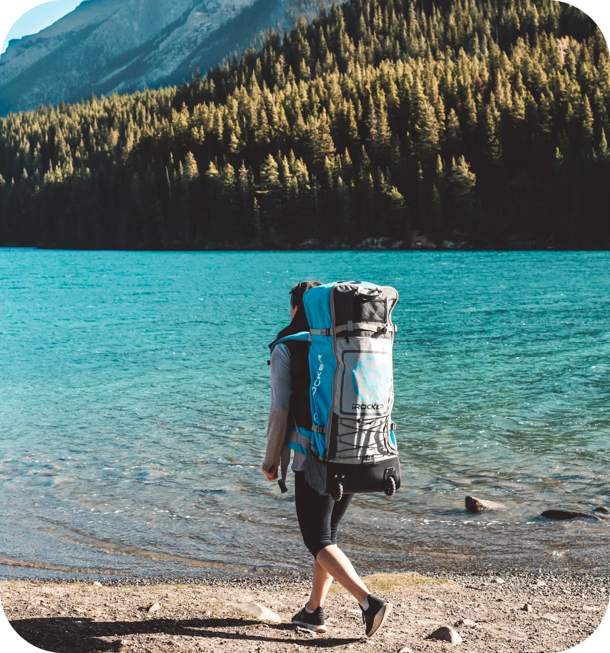 iROCKER lifestyle photo of a person carrying a paddle board backpack along a mountain lake shore.
