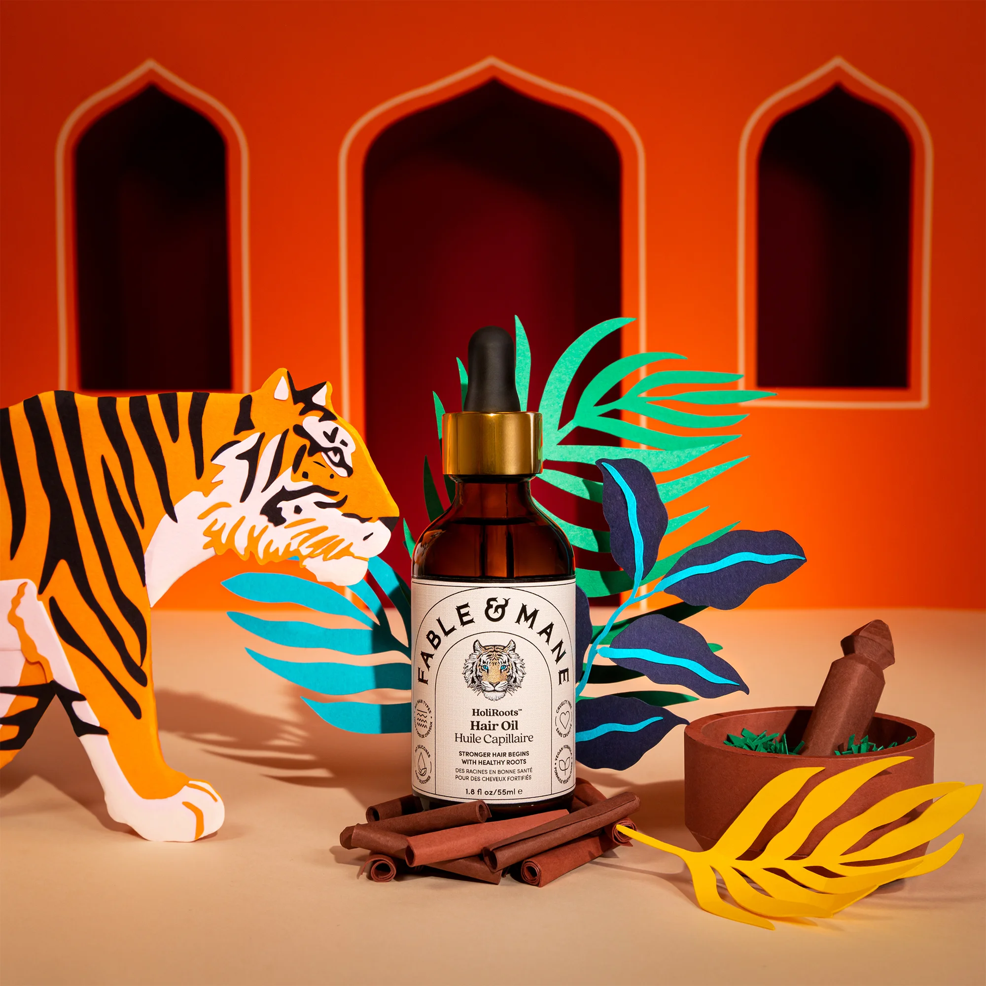 Fable & Mane HoliRoots hair oil bottle among paper-craft tiger, palm leaves, and cinnamon sticks.