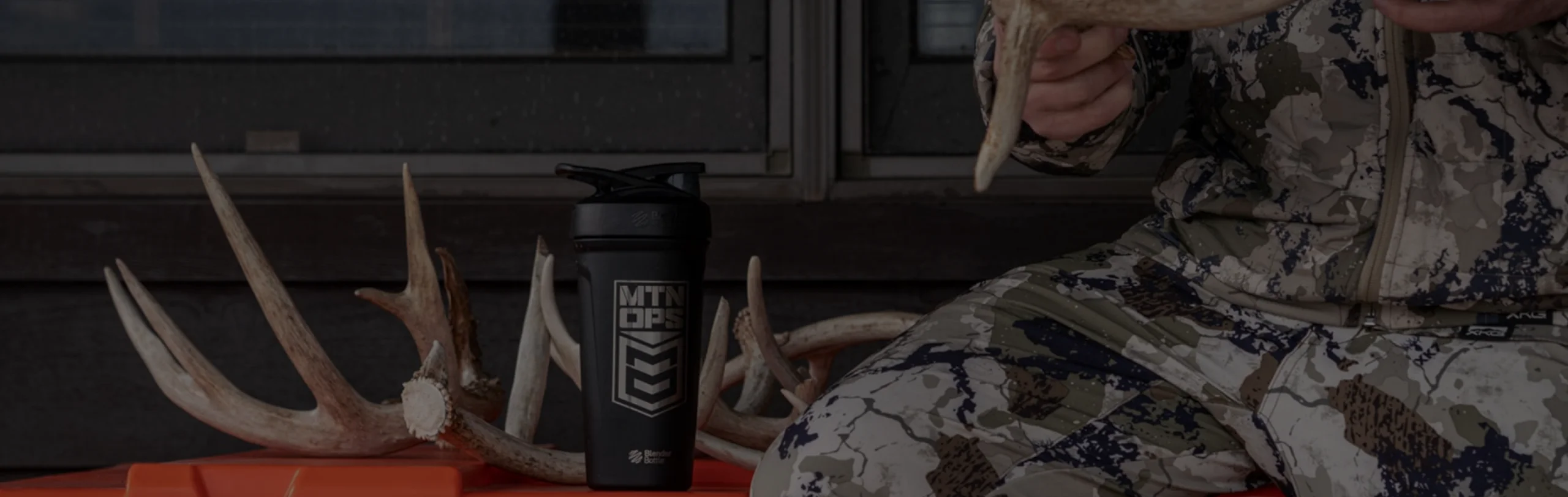 MTN OPS shaker bottle beside deer antlers with a hunter in camouflage holding a rack.