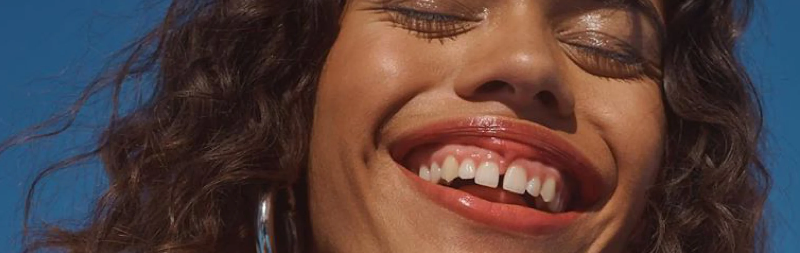 Close-up of a smiling woman with glossy curly hair against a blue sky for Briogeo hair care.