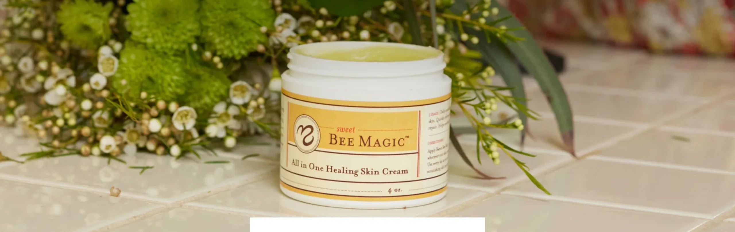 Open jar of Sweet Bee Magic healing skin cream on a tiled counter beside white flowers and greenery.