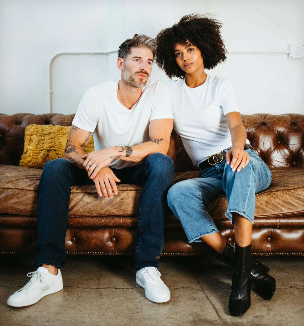 Fidelity Denim lifestyle photo of two models in jeans and white tees on a leather sofa.