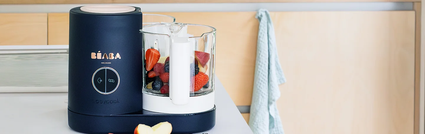 Beaba Babycook baby food maker filled with fresh berries beside apple slices on a kitchen counter.