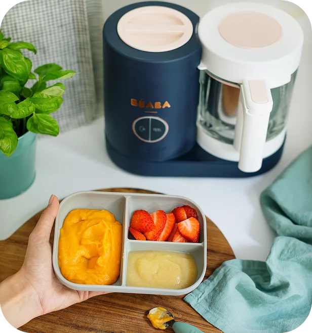 Béaba lifestyle photo of a baby food maker beside a tray of fresh fruit purees and strawberries.