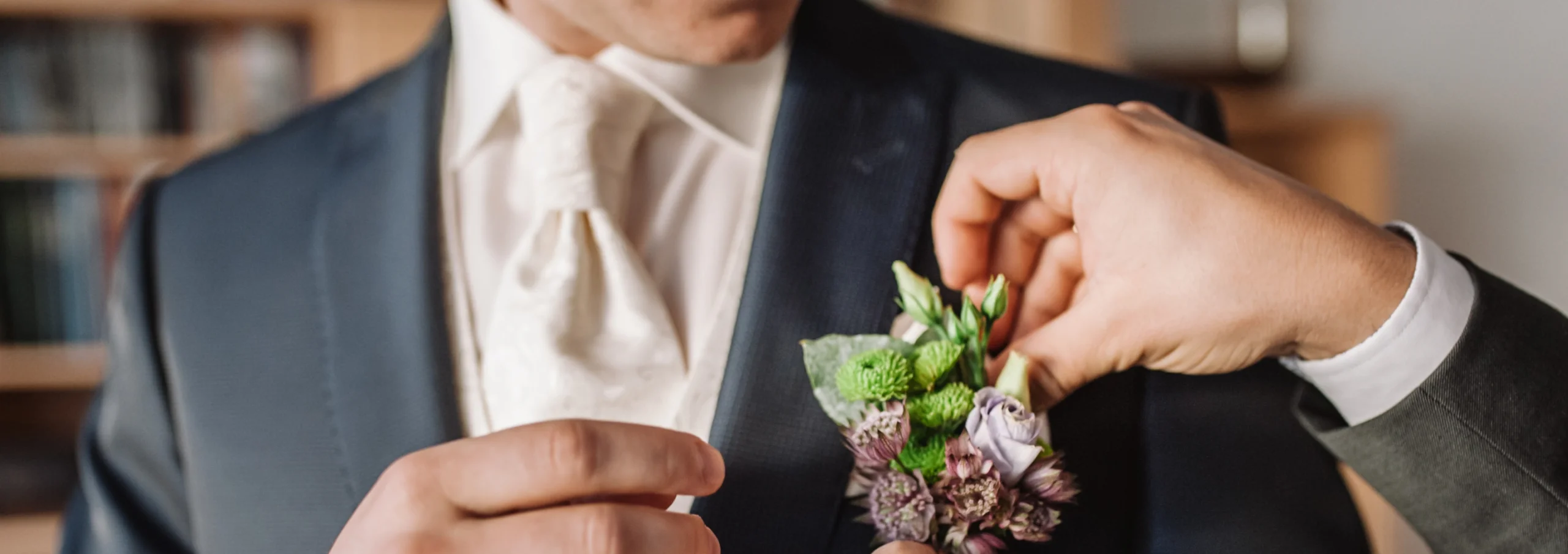 Hands pinning a floral boutonniere to the lapel of a groom's navy suit.
