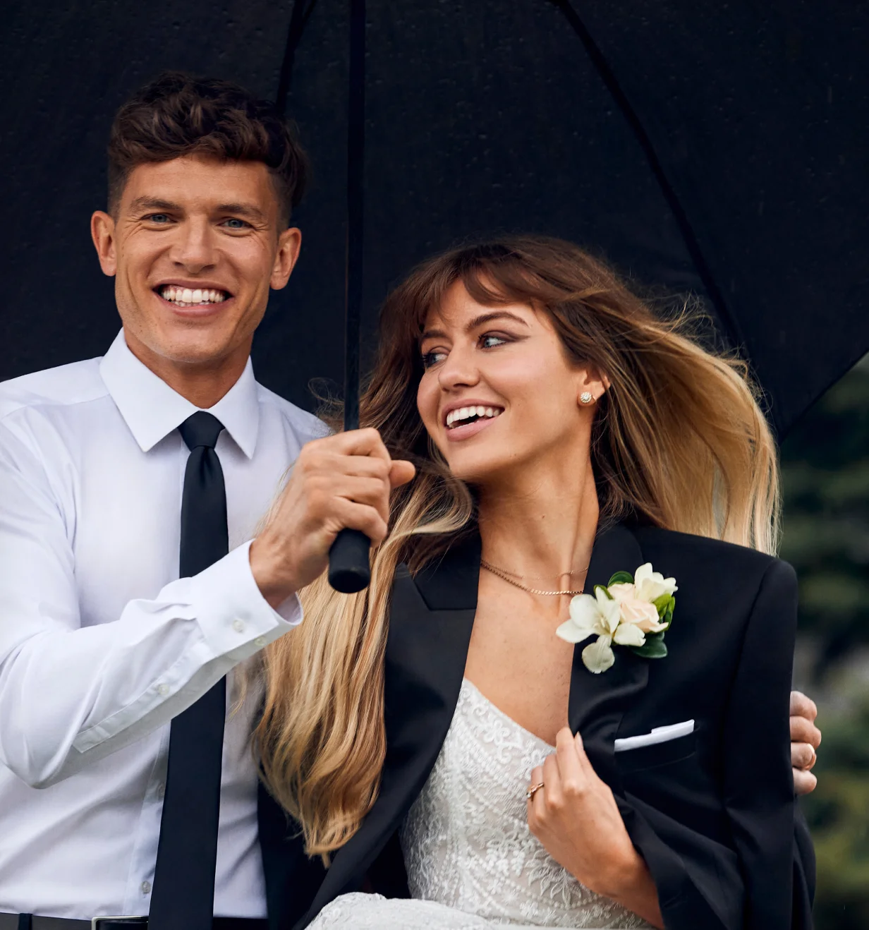 Grafton Apparel lifestyle photo of a smiling wedding couple in formalwear under a black umbrella.