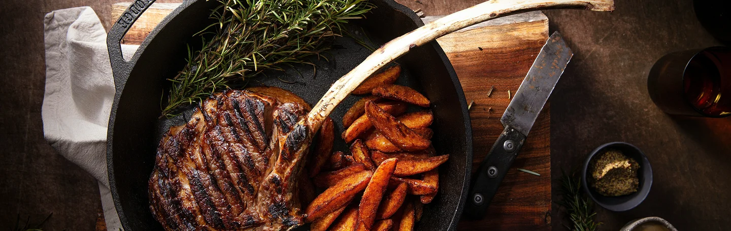 Grilled tomahawk steak and potato wedges in a cast-iron skillet with fresh rosemary.