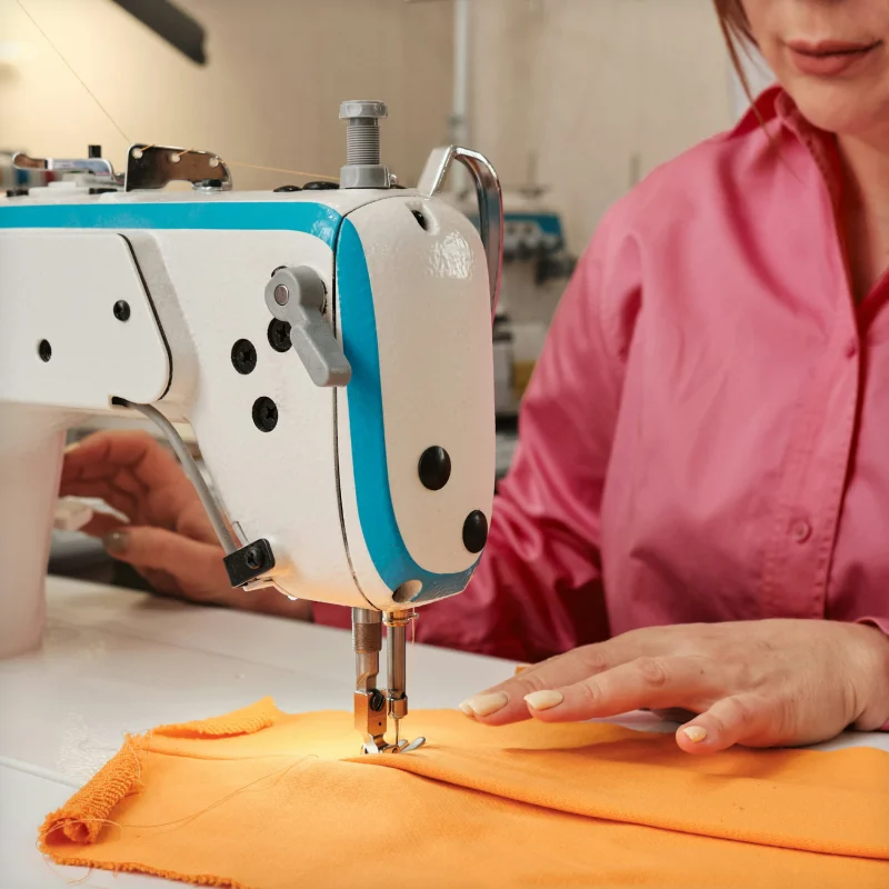 Worker in a pink shirt guiding orange fabric through an industrial sewing machine.