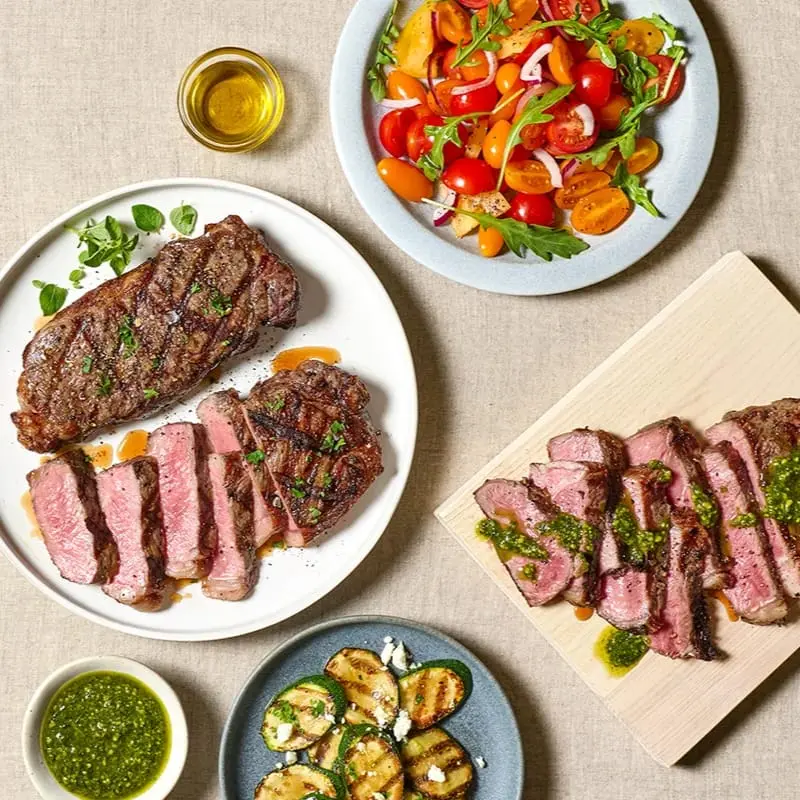 Overhead spread of grilled sliced steak with chimichurri, a tomato and arugula salad, and grilled zucchini on a linen table.