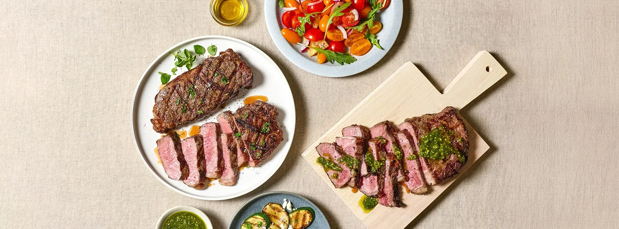 Grilled and sliced steaks with chimichurri alongside a tomato and arugula salad and grilled zucchini on a linen table.