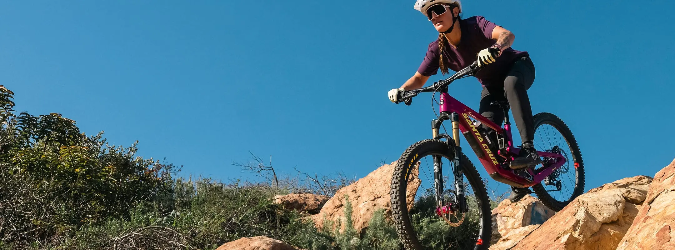 Mountain biker in a purple jersey riding down a rocky desert trail under a clear blue sky.