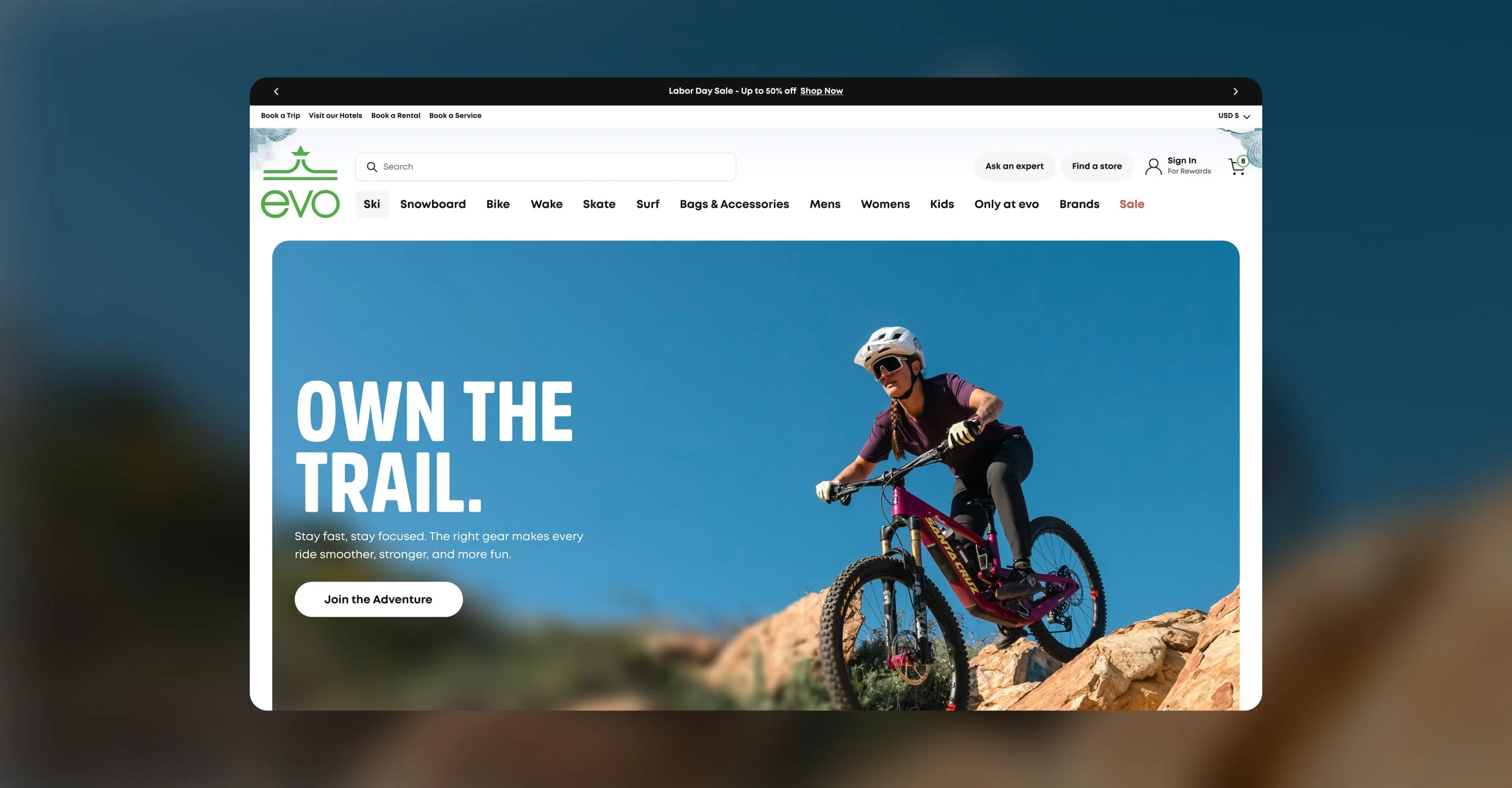 evo homepage on desktop with an Own the Trail hero banner and ski, snowboard, bike, and surf navigation.
