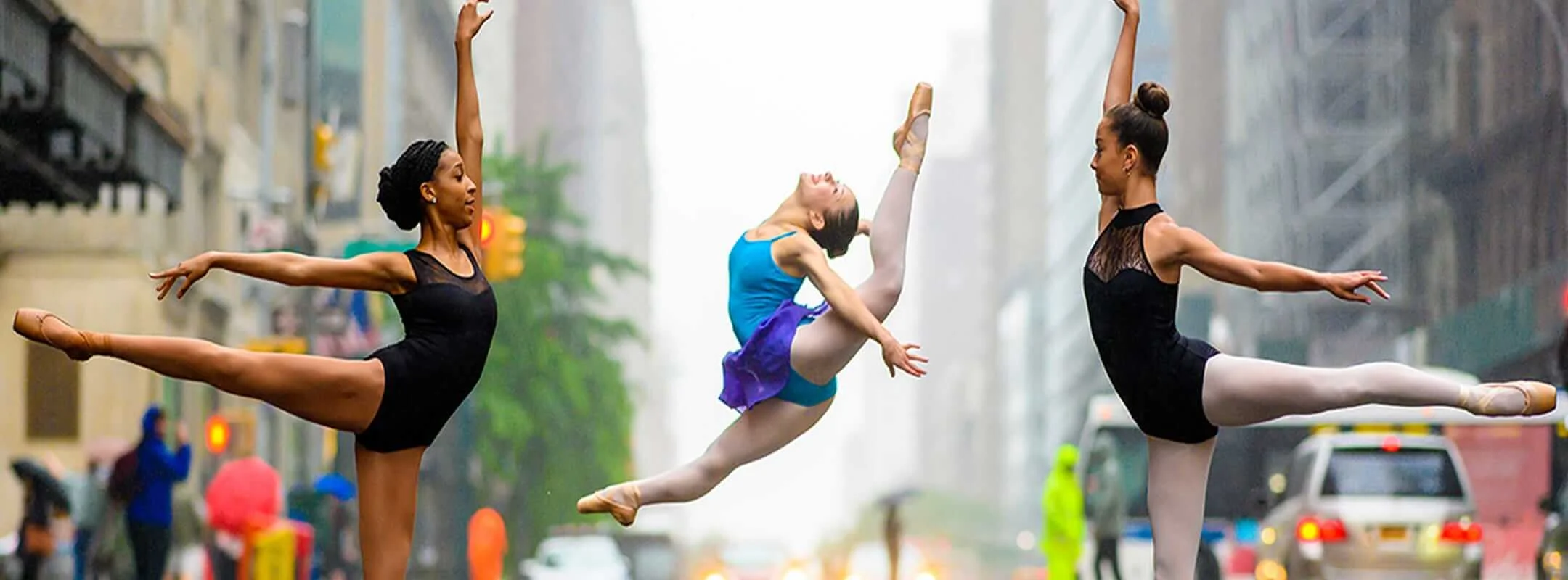 Three ballet dancers leaping and posing en pointe on a rainy city street.
