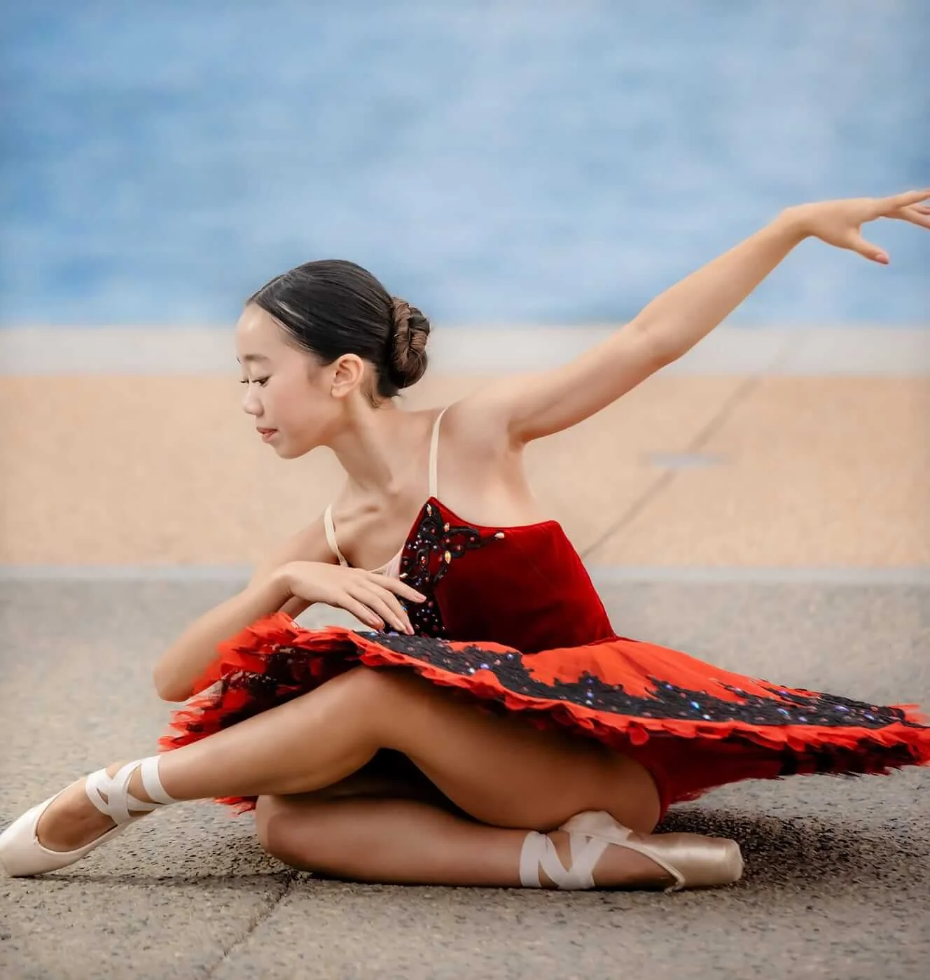 Young ballerina in a red and black tutu and pointe shoes posing seated on a waterfront promenade.