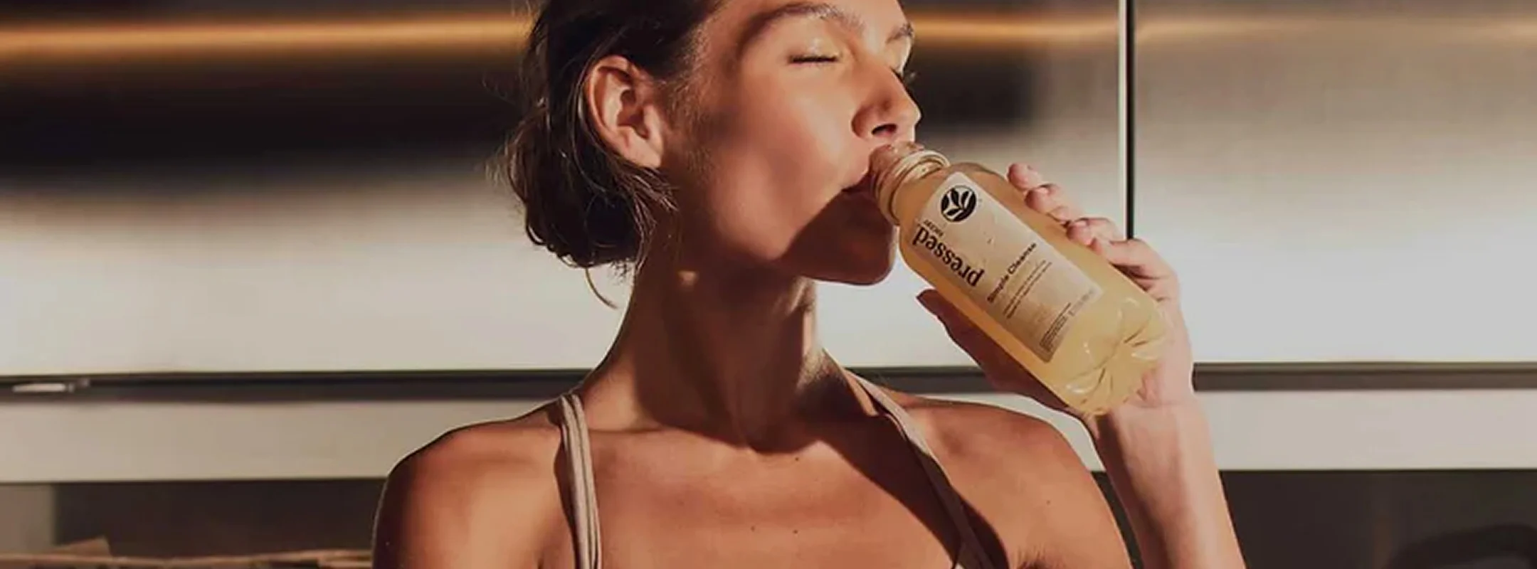 Woman with eyes closed sipping a bottle of Pressed juice in a sunlit stainless-steel kitchen.