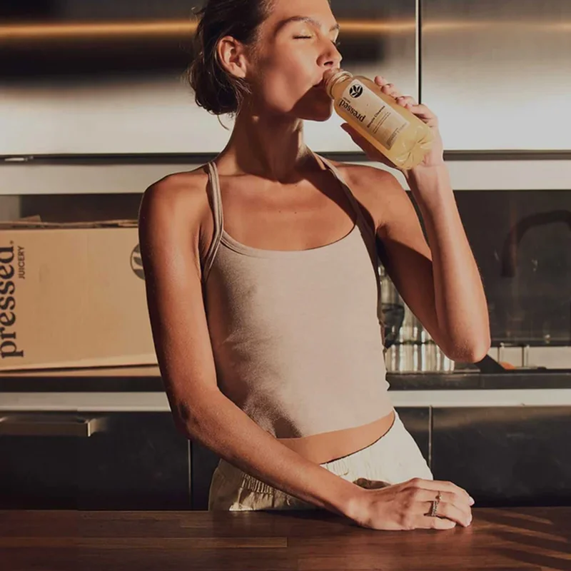 Woman in a neutral tank top drinking a bottle of Pressed Simple Cleanse juice at a kitchen counter.